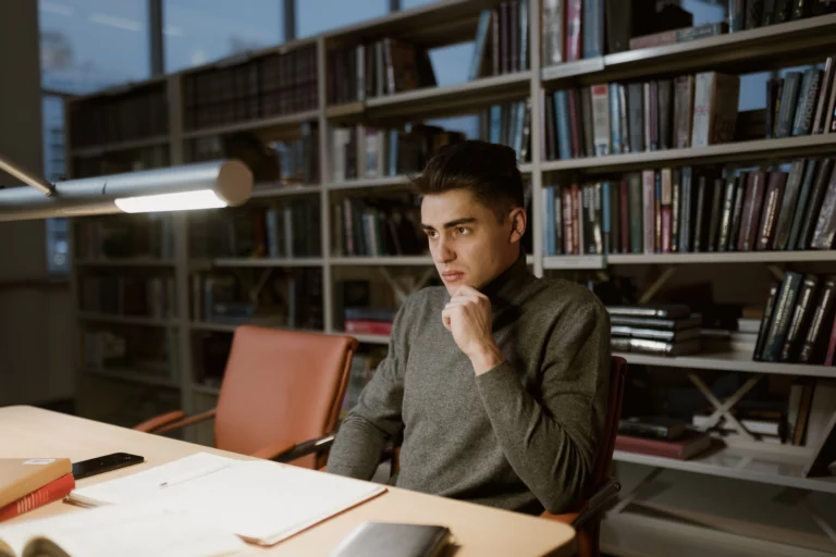 Jeune homme pensif assis à un bureau en bibliothèque, illustrant la peur de l'échec chez les étudiants